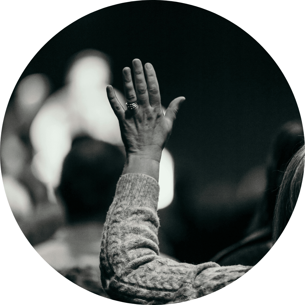 Black and white photo of a hand raised in a group setting, symbolizing a person asking a question or engaging in worship or teaching.