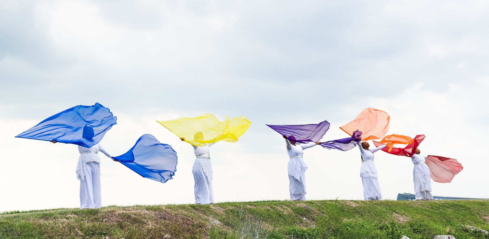 A group of women worshipping outdoors with colorful praise dance flags from Catch the Fire Worship Flags in blue, yellow, purple, orange, and red, dressed in flowing white garments. A vibrant expression of unity and worship movement under an open sky.