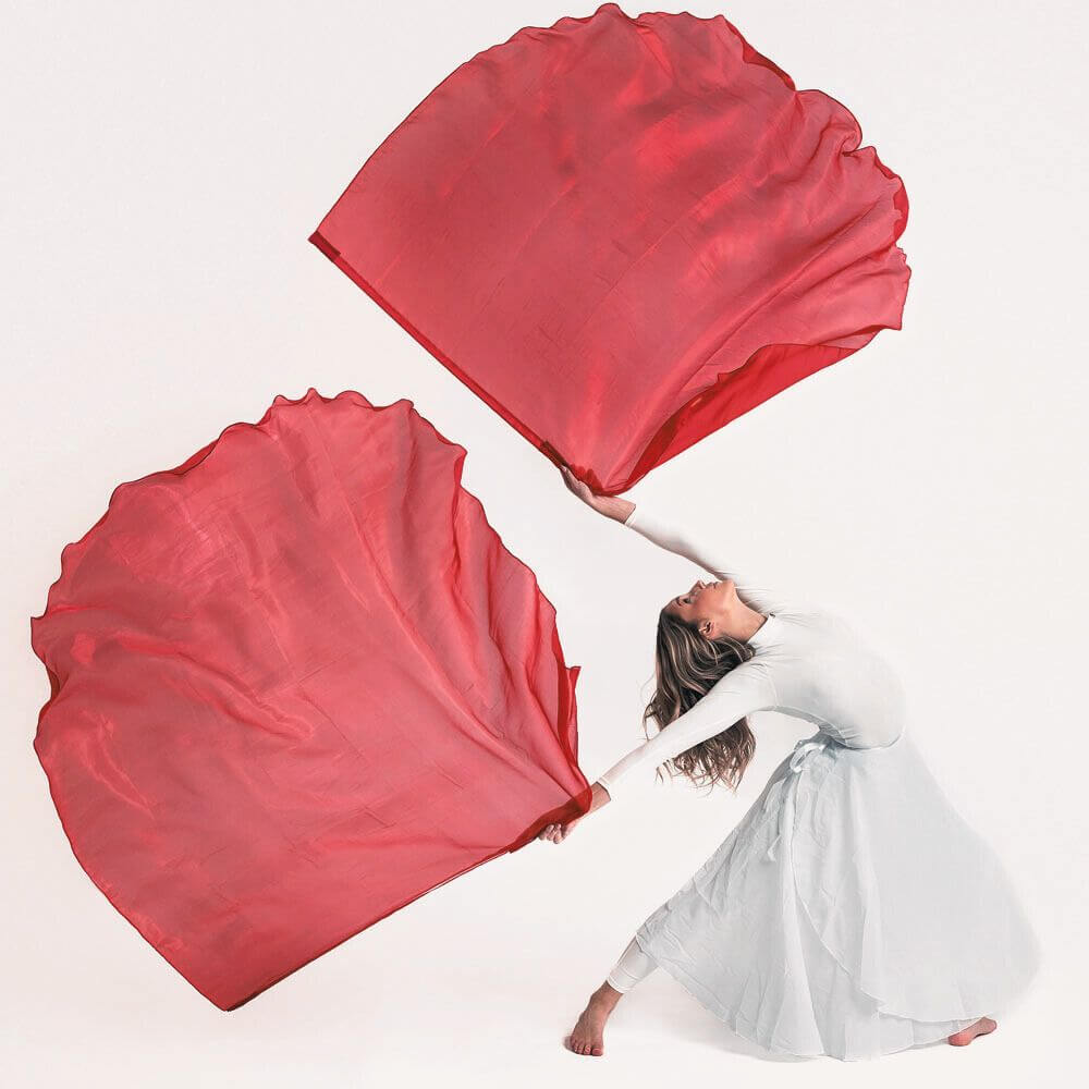 Worship dancer in a white dress gracefully extends red silk flags in a dramatic pose against a white background, expressing praise through movement