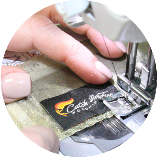 Close-up of hands sewing a Catch the Fire Worship Flags label onto a olive green sheer flag using a sewing machine, highlighting handcrafted quality and Christian artisan craftsmanship.