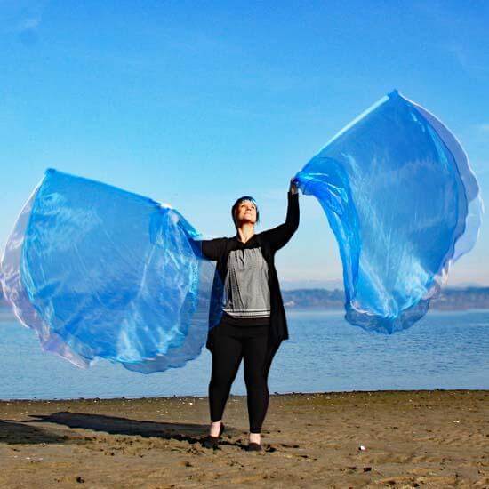 Woman worshipping with large blue and white sheer worship flags on a sandy beach, with a bright blue sky and calm water in the background.