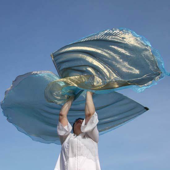 Person dressed in white worship attire raises a pair of flowing worship flags against a clear blue sky. The flags are made of sheer fabric with olive green and light blue hues, shimmering in the sunlight as they ripple through the air.